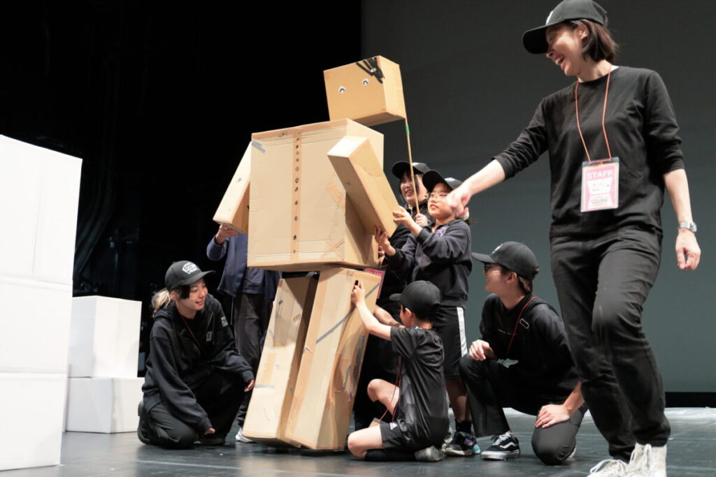 ダンボール箱を持った子どもたちが集まって、一つのキャラクターを作って動かしている