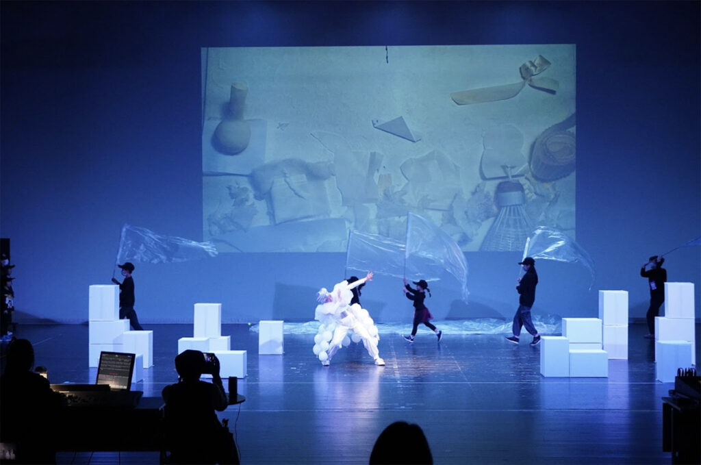 出演者や小道具を持った子どもたちが舞台上にいる。背景には綿・リボン・バドミントンのシャトルなどが置かれている写真が投影されている。